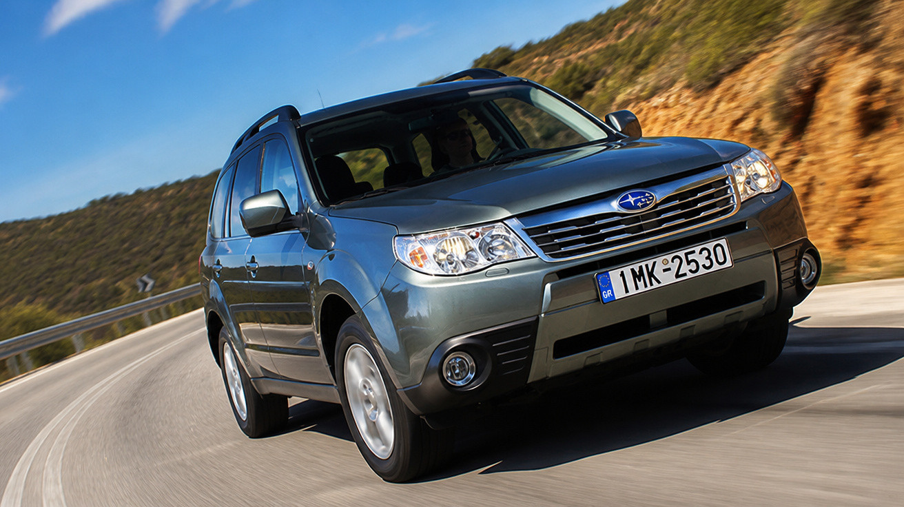 Carro SUV cinza da marca Subaru em estrada curvada com vegetação e morros ao fundo.