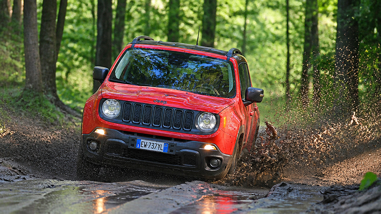 Jeep vermelho off-road atravessando lama em estrada cercada por floresta verde.