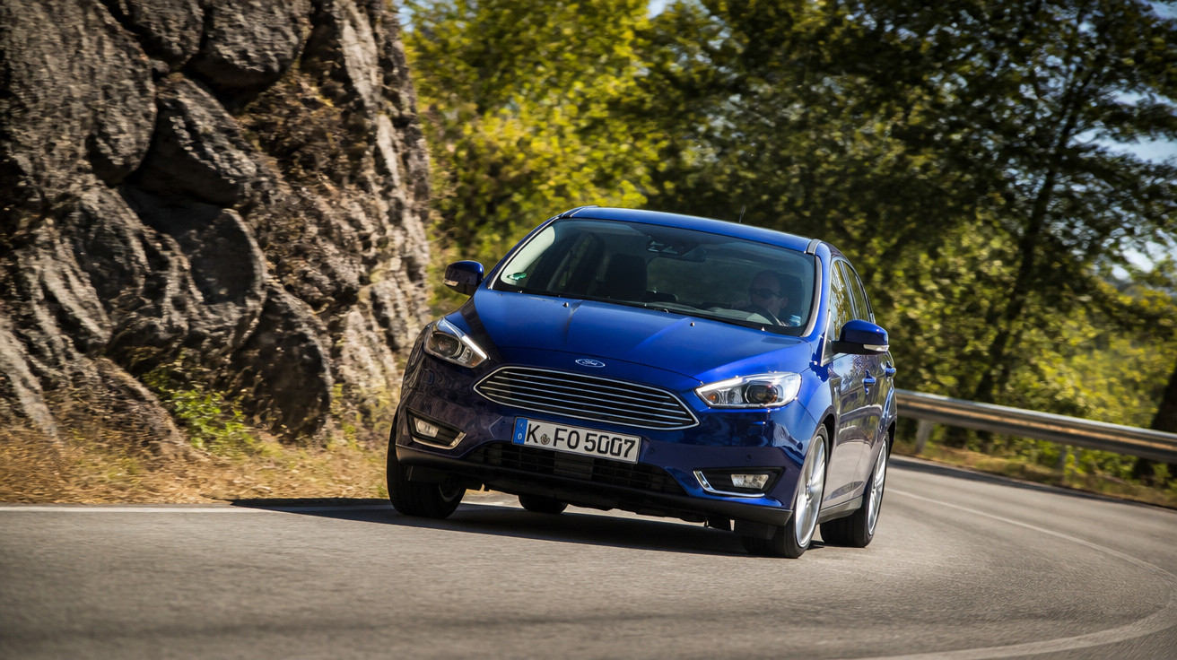 Carro azul modelo Ford Focus fazendo curva em estrada cercada por árvores e rochas.