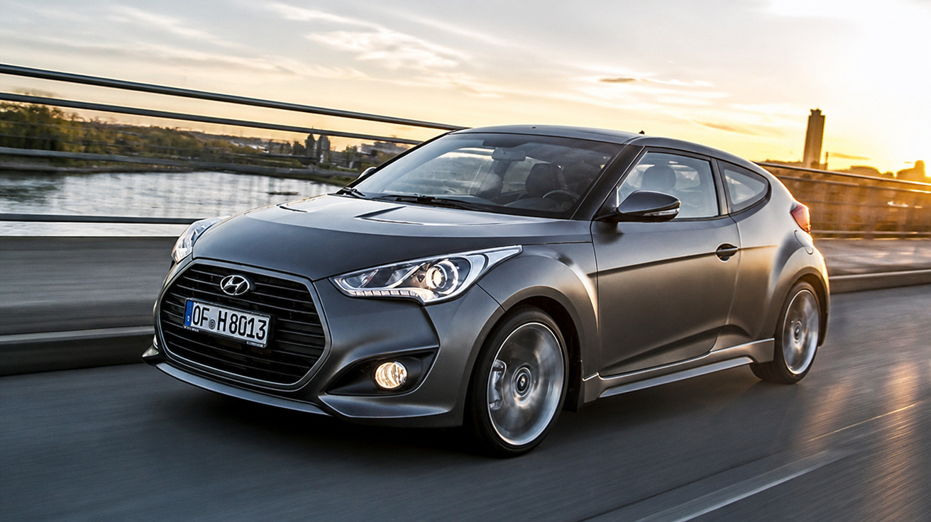 Carro esportivo Hyundai cinza escuro em movimento na estrada durante o pôr do sol.