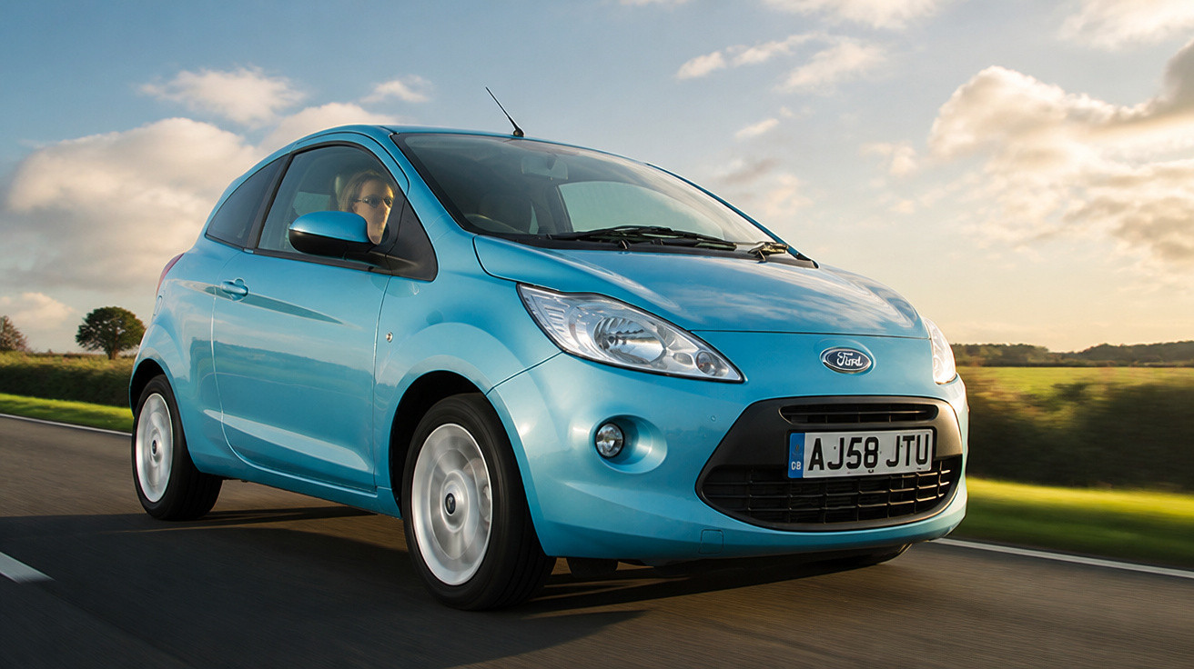 Carro Ford hatch compacto azul dirigindo em estrada sob céu parcialmente nublado ao entardecer.